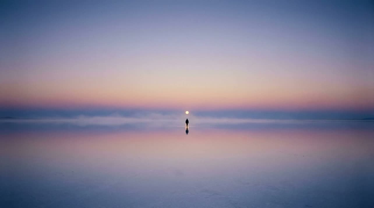 A lone figure crossing a mirror-smooth salt flat toward a low sun on the horizon, at the hour between night and dawn.